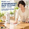 A woman in a cream cardigan wipes breakfast crumbs from a wooden table with a white fruit-print cloth beside a matching printed roll.